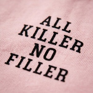 May include: Close-up of a light pink fabric with the phrase "ALL KILLER NO FILLER" printed in bold black letters. The text is slightly angled, creating a dynamic visual effect. The fabric's texture is visible.