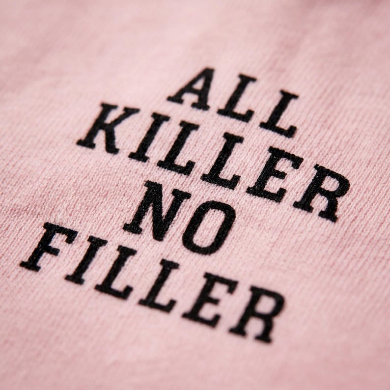 May include: Close-up of a light pink fabric with the phrase "ALL KILLER NO FILLER" printed in bold black letters. The text is slightly angled, creating a dynamic visual effect. The fabric's texture is visible.