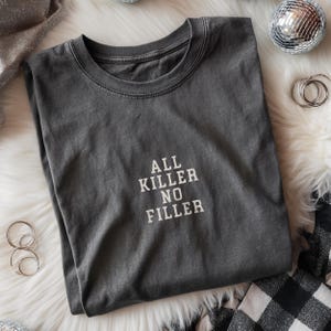 May include: A dark gray t-shirt with the text "ALL KILLER NO FILLER" in white block letters. The shirt is folded and resting on a white, fluffy surface. Decorative silver items are in the background.
