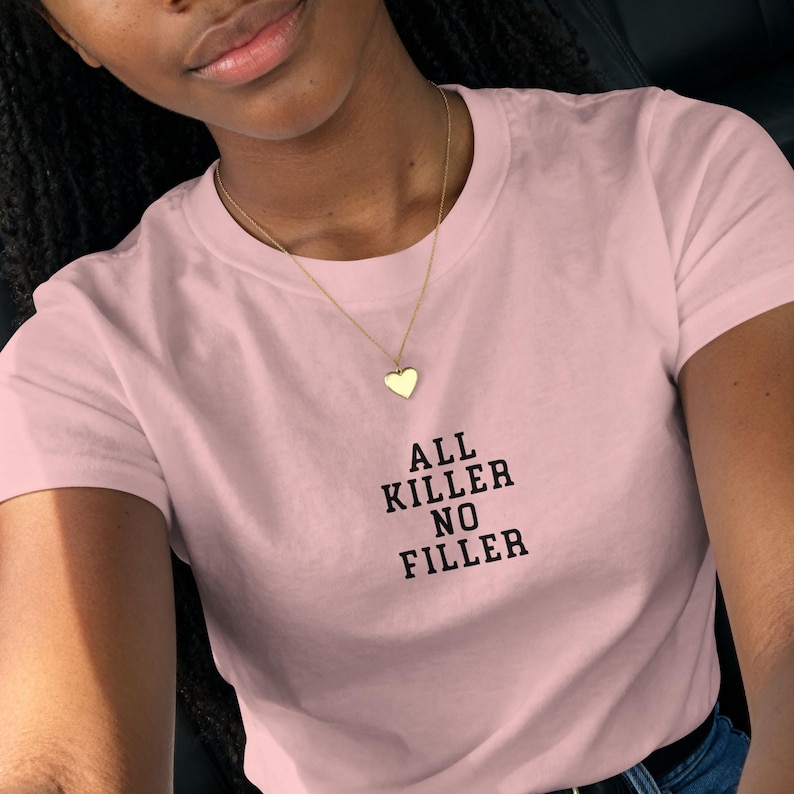 May include: A light pink t-shirt with the text "ALL KILLER NO FILLER" in black. A gold heart pendant hangs from a delicate gold chain. The shirt is a crew neck style.