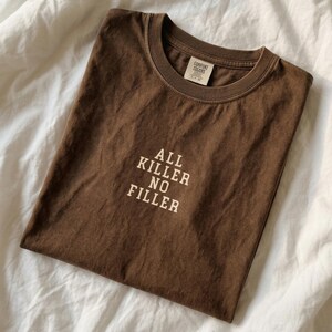 May include: A brown t-shirt with the text "ALL KILLER NO FILLER" in white block letters. The shirt is folded and has a tag that reads "COMFORT COLORS". The shirt is made of a soft material.