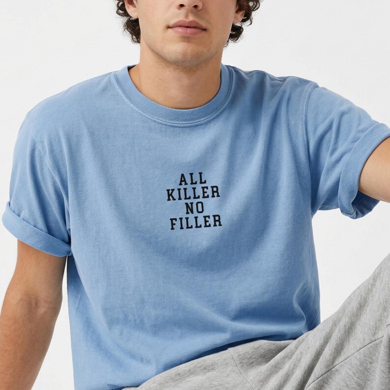 May include: A light blue t-shirt with the text "ALL KILLER NO FILLER" in black. The short-sleeved shirt is made of a soft material and has rolled-up sleeves. The shirt is a casual style.