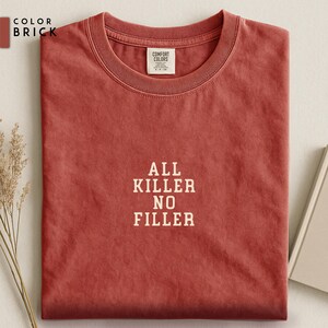 May include: Brick-colored t-shirt with the text "ALL KILLER NO FILLER" in white. The shirt is folded neatly, showcasing the crew neck and the Comfort Colors label. The fabric has a slightly textured appearance.