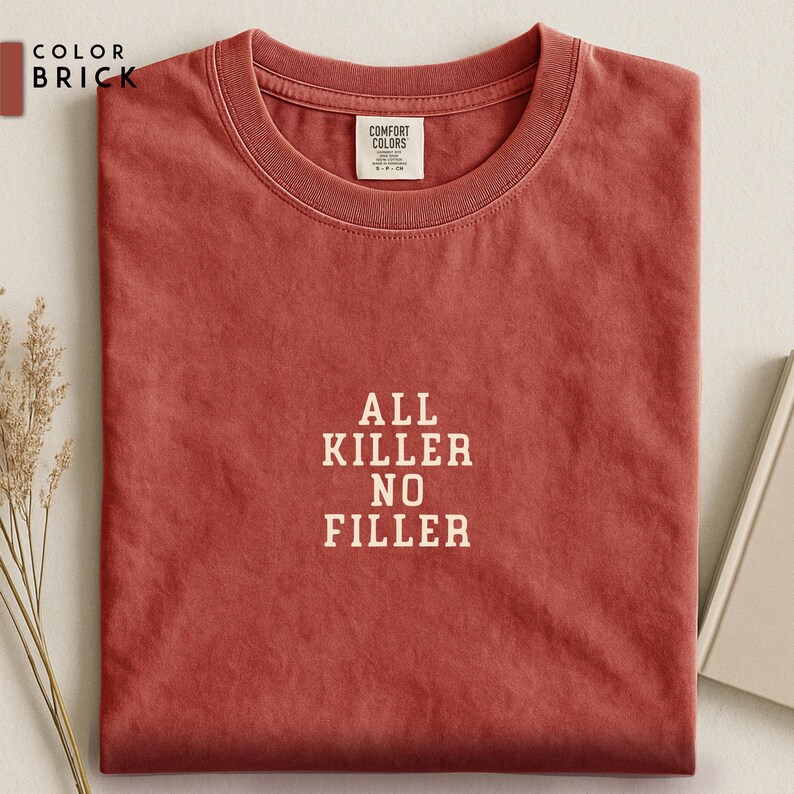 May include: Brick-colored t-shirt with the text "ALL KILLER NO FILLER" in white. The shirt is folded neatly, showcasing the crew neck and the Comfort Colors label. The fabric has a slightly textured appearance.