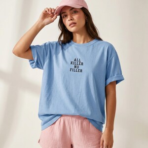 May include: A light blue t-shirt with the text "ALL KILLER NO FILLER" in black. The shirt has rolled-up sleeves. The person is wearing a pink baseball cap and pink shorts.