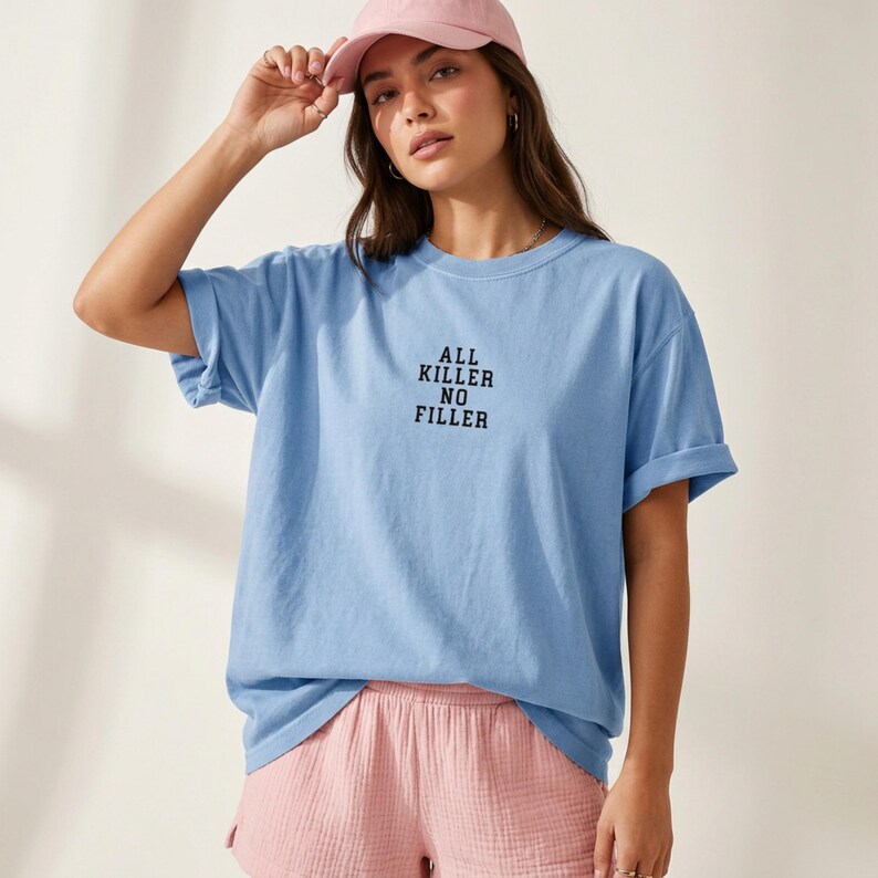 May include: A light blue t-shirt with the text "ALL KILLER NO FILLER" in black. The shirt has rolled-up sleeves. The person is wearing a pink baseball cap and pink shorts.