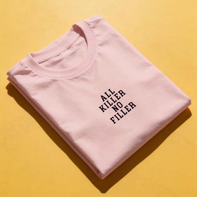 May include: A light pink t-shirt with the text "ALL KILLER NO FILLER" printed in black. The shirt is folded neatly on a yellow surface. The t-shirt has a crew neck.