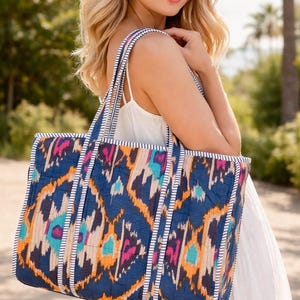 May include: A large tote bag with a vibrant, patterned design. The bag features a navy blue background with an ikat-style pattern in orange, pink, and turquoise. White and blue striped trim accents the edges and straps. The bag is carried over the shoulder.