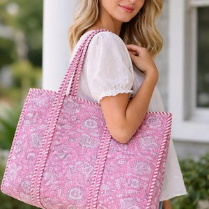 May include: A pink tote bag with a floral design. The bag features white and pink rope handles and trim. The bag is held by a person wearing a white top and blue jeans.