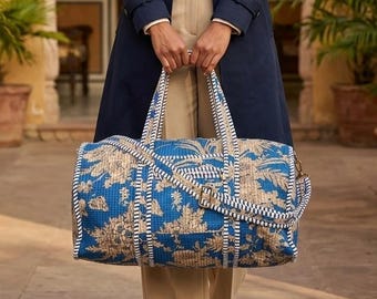 Könnte beinhalten: Eine Frau in einem marineblauen Trenchcoat hält eine blau-beige gemusterte Reisetasche. Die Tasche hat ein Blumenmuster und weiße Verzierungen. Sie trägt eine cremefarbene Bluse, beige Hose und weiße Loafer. Im Hintergrund sind Palmen und ein gelbes Gebäude zu sehen.