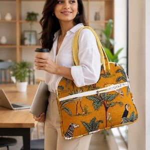 Op de afbeelding: Een gewatteerde tote bag van mosterdgele stof met een gedetailleerde jungle-print met diverse dieren en tropische planten. De tas is voorzien van een wit voorvak met rits, stevige gele hengsels met een patroon en een witte afwerking.