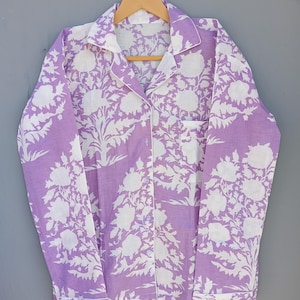 May include: A lavender pajama top with a white floral design. The long-sleeved shirt features a button-down front, a chest pocket, and white trim on the collar, cuffs, and pocket. It is displayed on a wooden hanger.