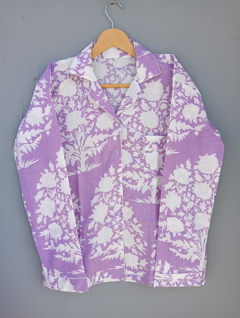 May include: A lavender pajama top with a white floral design. The long-sleeved shirt features a button-down front, a chest pocket, and white trim on the collar, cuffs, and pocket. It is displayed on a wooden hanger.