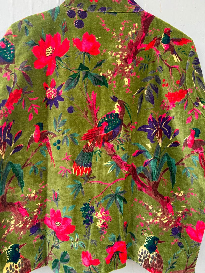 Op de afbeelding: Een jas met lange mouwen van olijfgroen fluweel met een levendige botanische print. Het patroon toont diverse kleurrijke vogels op takken, afgewisseld met felrode bloemen, paarse bloesems en gele florale accenten.