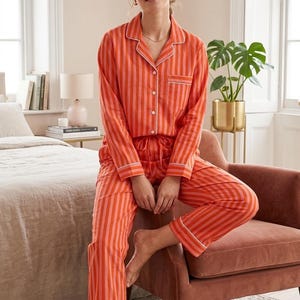May include: An orange and pink striped pyjama set. The long-sleeved top has a button-down front and white trim. The matching trousers have a drawstring waist and white trim. The pyjamas are made of a soft fabric.