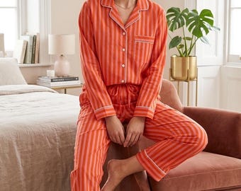 May include: An orange and pink striped pyjama set. The long-sleeved top has a button-down front and white trim. The matching trousers have a drawstring waist and white trim. The pyjamas are made of a soft fabric.