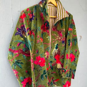 Op de afbeelding: Een jas met lange mouwen van olijfgroene fluwelen stof met een levendige print van rode bloemen, groene bladeren en kleurrijke vogels. De binnenvoering is zichtbaar bij de kraag en de opening aan de voorzijde, met een wit en bruin verticaal streeppatroon.