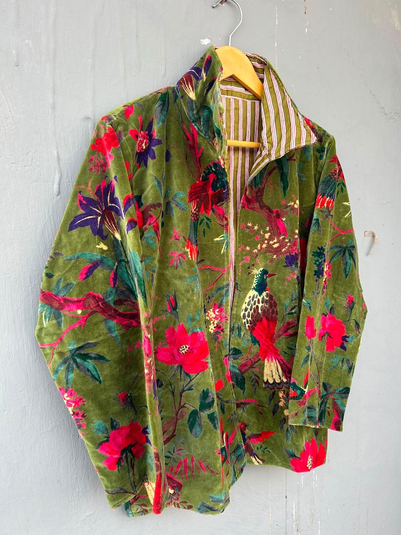 Op de afbeelding: Een jas met lange mouwen van olijfgroene fluwelen stof met een levendige print van rode bloemen, groene bladeren en kleurrijke vogels. De binnenvoering is zichtbaar bij de kraag en de opening aan de voorzijde, met een wit en bruin verticaal streeppatroon.