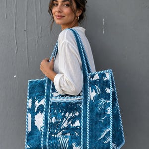 May include: A large tote bag with a blue and white tropical palm tree pattern. The bag has a rectangular shape with a pocket on the front and blue rope handles. The bag is being carried by a person.