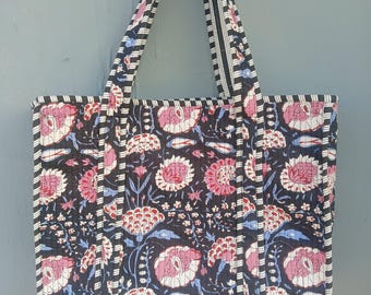 Puede incluir: Un bolso tote acolchado con un estampado floral. El bolso presenta un fondo oscuro con diseños florales rosas, blancos y azules. Las asas y los bordes están adornados con rayas blancas y negras. El bolso está diseñado para transportar objetos.
