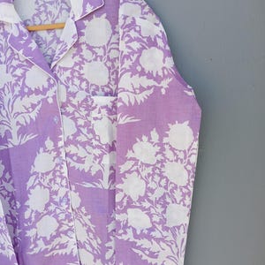 May include: A lavender pyjama set featuring a white floral design. The button-down shirt has a chest pocket and white trim. The fabric is hanging on a wooden hanger against a grey background.