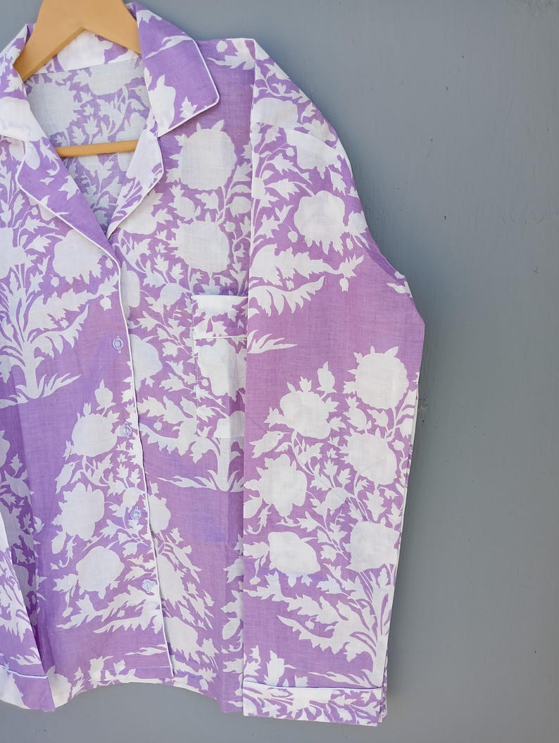 May include: A lavender pyjama set featuring a white floral design. The button-down shirt has a chest pocket and white trim. The fabric is hanging on a wooden hanger against a grey background.