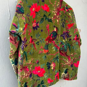 Op de afbeelding: Een jas met lange mouwen van olijfgroen fluweel met een levendige botanische print. De stof toont felrode bloemen, paarse accenten en gedetailleerde illustraties van vogels die op takken zitten over het gehele oppervlak van het kledingstuk.
