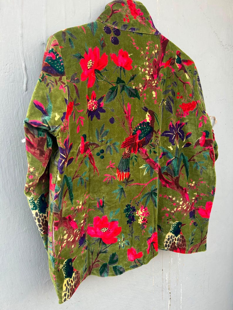 Op de afbeelding: Een jas met lange mouwen van olijfgroen fluweel met een levendige botanische print. De stof toont felrode bloemen, paarse accenten en gedetailleerde illustraties van vogels die op takken zitten over het gehele oppervlak van het kledingstuk.