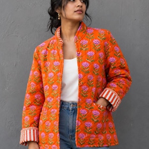 May include: A vibrant orange quilted jacket featuring a floral design in pink and green. The jacket has a stand-up collar, two pockets, and striped cuffs. The model is wearing blue jeans and a white top.