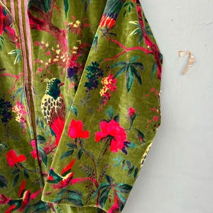 Op de afbeelding: Een jas met lange mouwen van olijfgroene fluwelen stof met een levendige botanische print met kleurrijke vogels, felroze bloemen en groen gebladerte. De binnenvoering is zichtbaar en toont een wit en rood verticaal gestreept patroon.