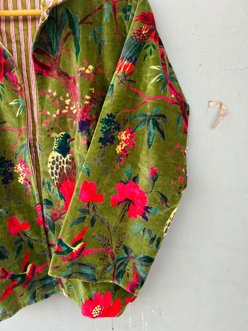 Op de afbeelding: Een jas met lange mouwen van olijfgroene fluwelen stof met een levendige botanische print met kleurrijke vogels, felroze bloemen en groen gebladerte. De binnenvoering is zichtbaar en toont een wit en rood verticaal gestreept patroon.