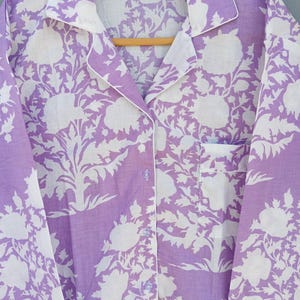 May include: A lavender pyjama set with a white floral pattern. The button-down shirt has a chest pocket and white trim along the collar, cuffs, and button placket. The pyjamas are hanging on a wooden hanger.