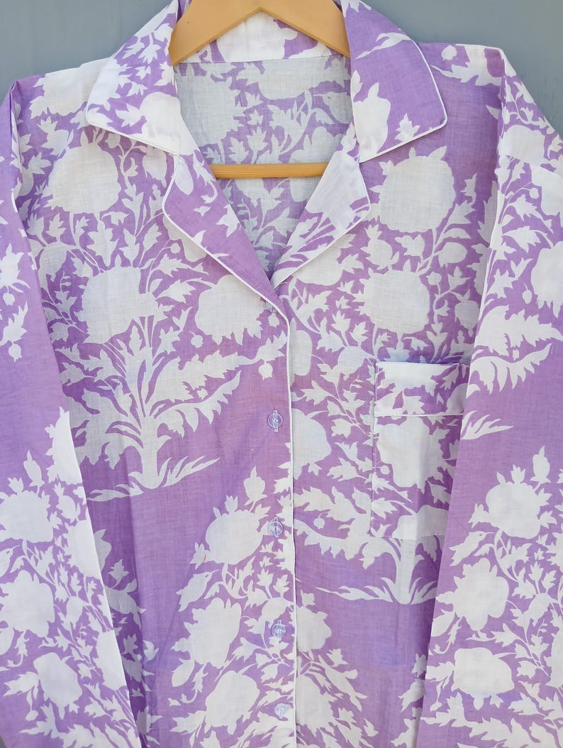 May include: A lavender pyjama set with a white floral pattern. The button-down shirt has a chest pocket and white trim along the collar, cuffs, and button placket. The pyjamas are hanging on a wooden hanger.