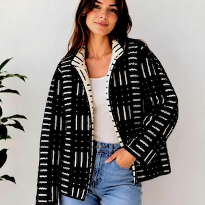 May include: A black jacket featuring a white geometric pattern of vertical lines and small dots. The jacket has a cream-coloured interior lining visible along the front opening and collar, worn over a white top and light blue denim jeans.