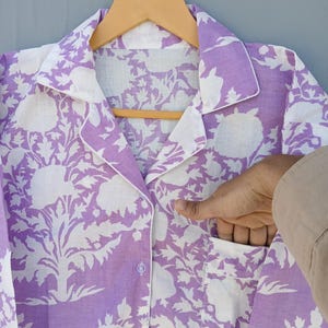May include: A lilac and white floral print pajama top with a notched collar and a pocket. The top is button-down and hangs on a wooden hanger. The floral pattern is a large, stylized design.
