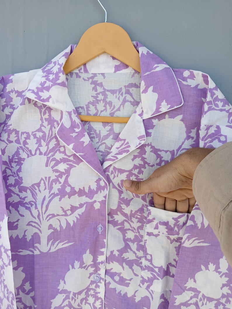 May include: A lilac and white floral print pajama top with a notched collar and a pocket. The top is button-down and hangs on a wooden hanger. The floral pattern is a large, stylized design.