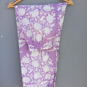 May include: Lavender and white floral print pajama trousers hanging on a wooden hanger. The trousers feature a repeating pattern of large white flowers and leaves against a lavender background. The trousers have an elasticated waistband and a drawstring.