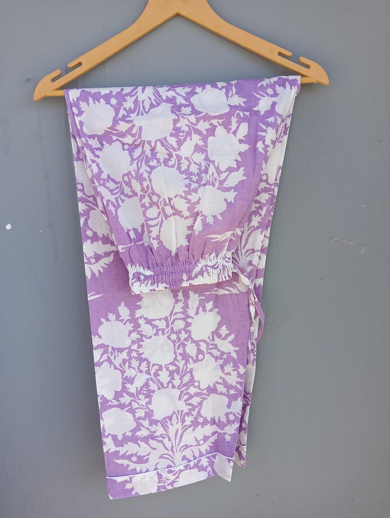 May include: Lavender and white floral print pajama trousers hanging on a wooden hanger. The trousers feature a repeating pattern of large white flowers and leaves against a lavender background. The trousers have an elasticated waistband and a drawstring.