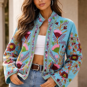 Könnte beinhalten: Hellblaue bestickte Jacke mit Stehkragen und langen Ärmeln. Die Jacke hat ein Blumenmuster in Rosa, Rot, Grün und Gelb. Die Jacke wird mit einem weißen Oberteil, einer blauen Jeans und einem braunen Gürtel getragen.
