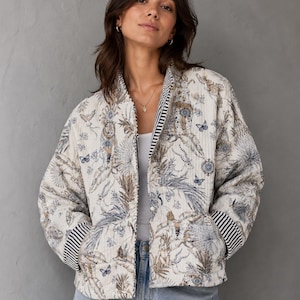 Può includere: Giacca trapuntata color crema con stampa botanica di foglie blu e marroni e farfalle. La giacca ha un bordo a righe sul colletto e sui polsini. La giacca è aperta, rivelando un top bianco e jeans blu.