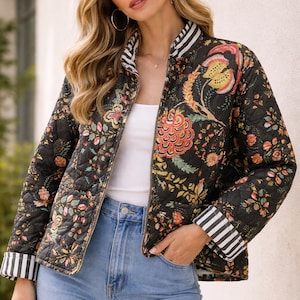 Op de afbeelding: Zwart gewatteerd jack met een levendig bloemmotief in oranje, geel en roze. De jas heeft een gestreepte kraag en manchetten en een ritssluiting. De jas wordt gedragen met een lichtblauwe jeans.