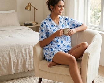 Puede incluir: Un conjunto de pijama de dos piezas compuesto por una camisa de manga corta con botones y pantalones cortos a juego, con un estampado floral azul sobre fondo blanco. La camisa tiene cuello de solapa y el pantalón corto un corte holgado.