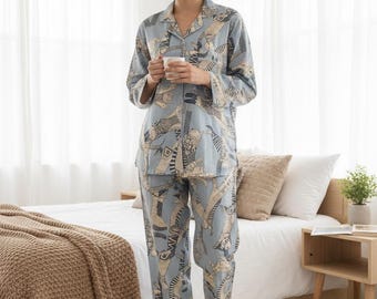 Puede incluir: Conjunto de pijama azul claro con un patrón repetido de figuras abstractas. El conjunto incluye una camisa con botones y pantalones a juego. La modelo sostiene una taza blanca. El pijama está hecho de una tela suave y ligera.