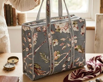 Puede incluir: Un bolso tote gris con un estampado floral, sostenido por una persona. El bolso presenta un diseño acolchado con detalles en blanco y rojo. El estampado floral incluye flores y hojas rosas, blancas y verdes. El bolso tiene dos asas largas y un cierre de cremallera.