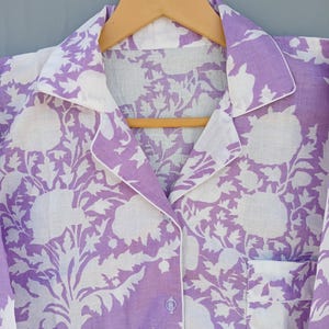 May include: A lilac pajama top with a white floral design and white trim. The button-down shirt features a chest pocket and is suspended on a wooden hanger. The floral pattern is a repeating design.