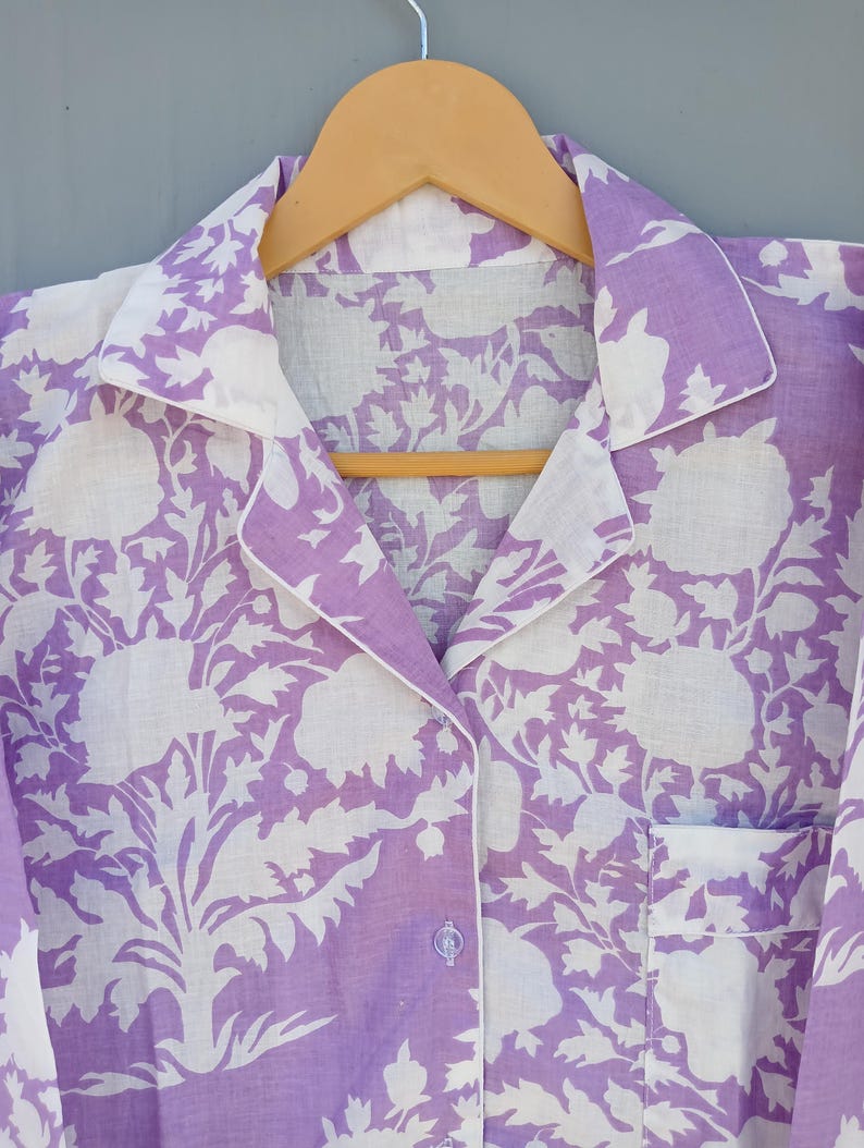 May include: A lilac pajama top with a white floral design and white trim. The button-down shirt features a chest pocket and is suspended on a wooden hanger. The floral pattern is a repeating design.