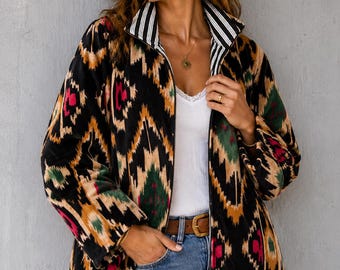 Op de afbeelding: Een jack met ritssluiting en een geometrisch patroon in zwart, beige, groen en roze, geïnspireerd op ikat. Het jack heeft een hoge kraag met een zwart-witte verticaal gestreepte voering, lange mouwen en een ritssluiting over de gehele voorzijde.