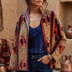 Puede incluir: Una chaqueta de manga larga con un patrón repetitivo de tigres sobre rayas verticales naranjas y azul marino. La chaqueta tiene un cuello alto y se lleva sobre una camiseta de tirantes azul marino y vaqueros azul oscuro. La tela tiene una textura aterciopelada.