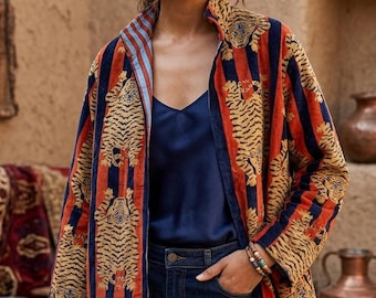 Puede incluir: Una chaqueta de manga larga con un patrón repetitivo de tigres sobre rayas verticales naranjas y azul marino. La chaqueta tiene un cuello alto y se lleva sobre una camiseta de tirantes azul marino y vaqueros azul oscuro. La tela tiene una textura aterciopelada.
