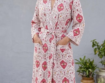 Puede incluir: Una bata de manga larga con base blanca y un patrón repetitivo de motivos en forma de diamante rosa que contienen ilustraciones de abejas, intercalados con pequeños detalles florales. La bata incluye un cinturón y dos bolsillos.